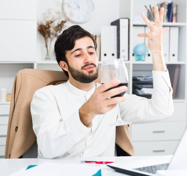 Positive young male manager with glass of wine having emotional online conversation at office desk - Foto, Imagem
