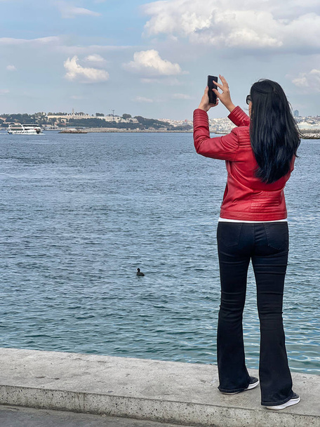 taking photo, back of young slim woman tourist taking photo of city view by sea with mobile or smartphone in Istanbul, Turkey. woman standing by sea with cellphone - Fotografie, Obrázek
