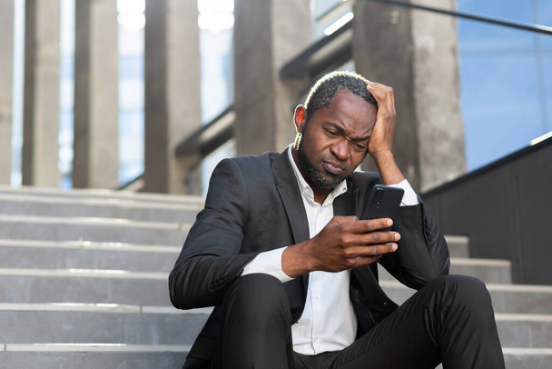 Sad african american businessman reading news in depression sitting on stairs outside office building worker holding smartphone. - Fotografie, Obrázek