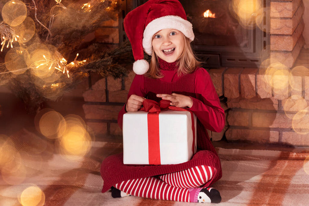 Karácsonyi otthon Kandalló atmoszféra. Kislány gyermek piros Mikulás kalap Szórakozás karácsonyi ajándék doboz közelében karácsonyfa - Fotó, kép