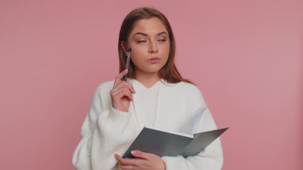 Thoughtful journalist woman with in white sweater making notes, writing down thoughts with pen into notepad notebook diary, to do list, good idea. Young adult girl isolated on pink studio background - Footage, Video