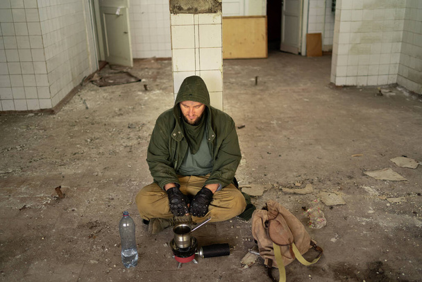 Постапокалипсис. Молодой человек сидит в заброшенном здании на полу, готовящем ужин на газовой горелке. концепция человека, путешествующего в пост-апокалиптическом мире в поисках пищи. копировальное пространство - Фото, изображение