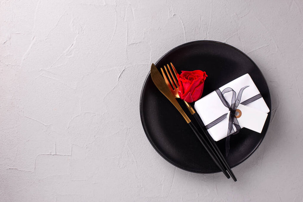 Romantic table setting. Black plate, knife, fork, box with present and empty tag  and red rose flower on textured grey background. Top view. Place fr text. Romantic postcard. - Foto, imagen