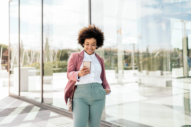 Happy black business woman using her mobile phone outdoors. She is wearing formal clothes and using earphones. - 写真・画像