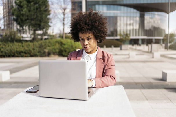 Pretty black business woman with afro hair using her laptop outdoors. She is wearing formal clothes. - Photo, Image