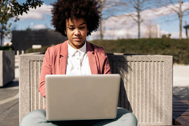 Business woman with afro hair wearing formal clothes sitting outdoors and using her computer. - Photo, Image