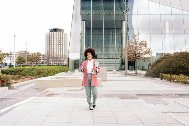 Happy black business woman walking in business area of the city and holding her laptop. She is drinking coffee too. - 写真・画像