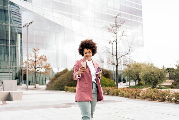 Happy black business woman using her mobile phone outdoors and holding coffee cup. She is wearing formal clothes and using earphones. - Foto, afbeelding