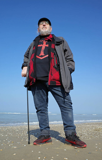 Un hombre mayor parado en la playa. Lleva una camisa con una cruz de templarios. Es hermoso día soleado en primavera. - Foto, imagen