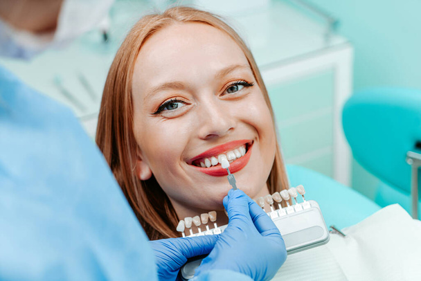 Attractive young woman with natural white teeth in dental clinic. Hands doctor dentist with teeth color palette next by face. Smile healthy teeth concept - Foto, Imagen