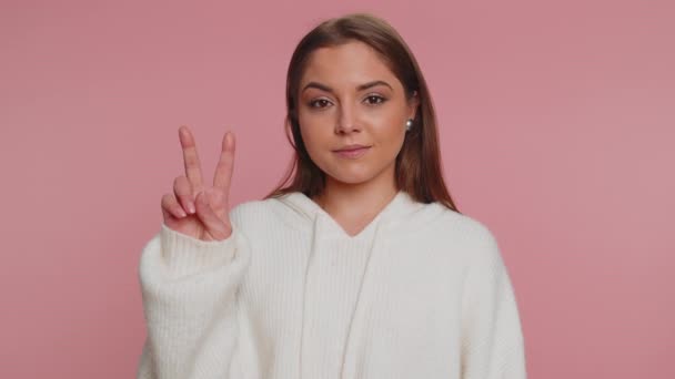 Hipster woman in white sweater showing victory v sign, hoping for success and win, doing peace gesture, smiling with kind optimistic expression. Young adult girl isolated on pink studio background - Footage, Video