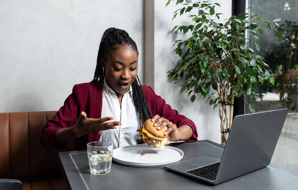 Young happy college exchange student or African American freelancer business woman having issue eating big fat tasty cheeseburger with melting cheese while she work on laptop computer in cafeteria on lunch break. - Fotografie, Obrázek