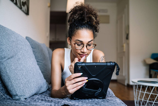 Afroameričanka v brýlích dotýkající se rtů a sledující video na tabletu, zatímco leží na gauči v obývacím pokoji doma - Fotografie, Obrázek