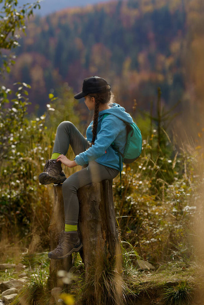 Une fille dans un sac à dos et une casquette est assise sur un tronc d'arbre dans les montagnes par une journée d'automne claire, regarde autour et ajuste une chaussette dans une chaussure. En arrière-plan, vous pouvez voir une forêt et un sommet de montagne. - Photo, image