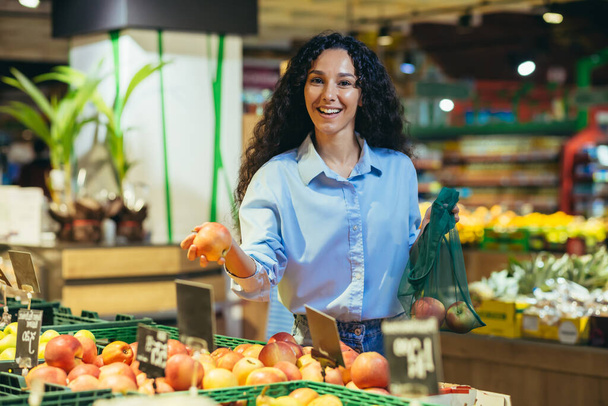 Портрет счастливой женщины-покупателя в супермаркете, латиноамериканка выбирает яблоки и фрукты улыбаясь и глядя в камеру, с корзиной продуктов выбирает товар. - Фото, изображение