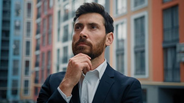 Caucasian smart puzzled thoughtful man hold hand to chin dreaming about career growth successful employer in stylish black suit stand outside modern office pondering idea planning business development - Fotó, kép