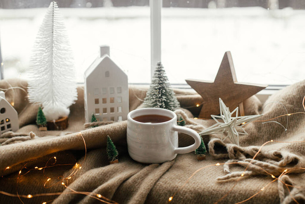 Kerststilleven, winterhygge thuis. Warme kop thee, kerstversiering, verlichting, huisje, ster op knusse deken op vensterbank. Gezellig thuis. Atmosferische schandalige stemming - Foto, afbeelding