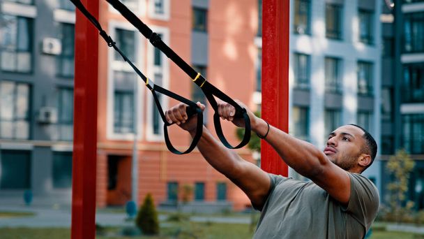 Izmos sportos erős latin-amerikai férfi testépítő sportoló használni sport felszerelést szabadtéri nyújtás edzés a város sportpálya lógó húzza up gyakorlat TRX hevederek aktív egészséges élet - Fotó, kép