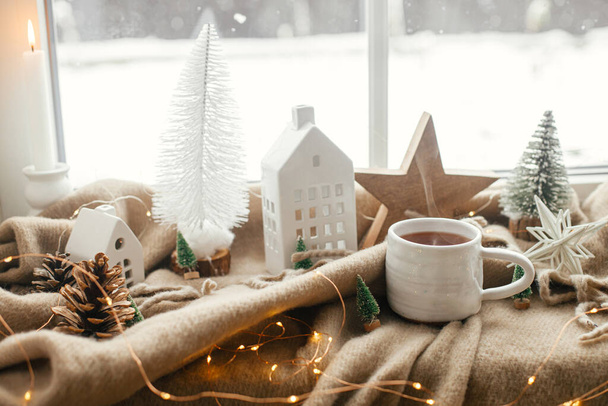 Kerststilleven, winterhygge thuis. Warme kop thee, kerstversiering, verlichting, huisje, ster op knusse deken op vensterbank. Gezellig thuis. Atmosferische schandalige stemming - Foto, afbeelding