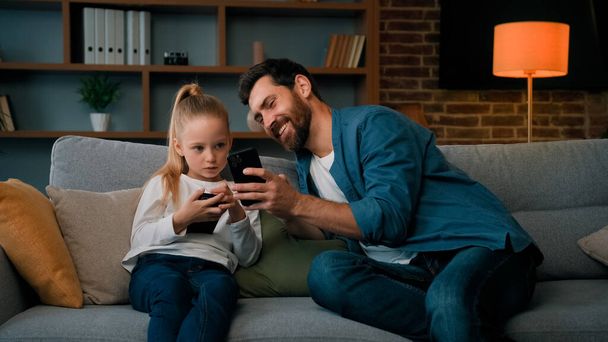 Caucasian man and little kid girl sit together at home hold modern phone browsing media content play games online adult dad teach child daughter use mobile apps showing funny video on wireless device - Photo, Image