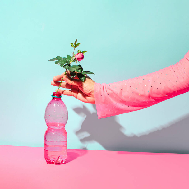 Mano femminile, bottiglia di plastica rosa e fiore di rosa, layout estetico floreale creativo contro la parete blu pastello.  - Foto, immagini
