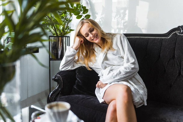 Portrait intérieur de jeune femme caucasienne aveugle maigre riant tout en étant assise sur un canapé noir luxueux à son salon d'esthéticienne SPA préféré. Peignoir blanc. Plantes et tasse à café. Photo de haute qualité - Photo, image