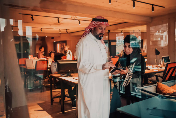 Portrait photo of Arab leader businessman with Muslim hijab woman discussing business projects while using tablets and smartphones in a modern glass office. - Photo, Image