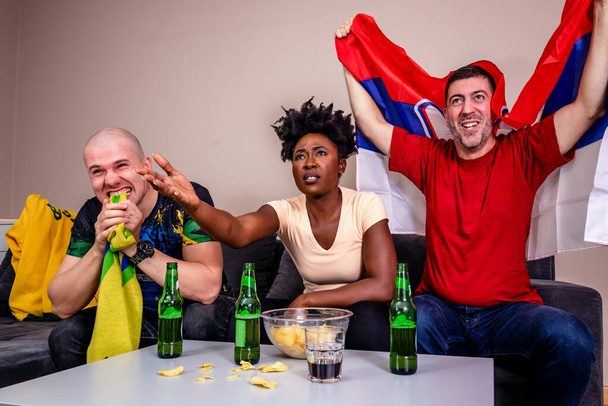 Φίλοι που παρακολουθούν ποδοσφαιρικό αγώνα στην τηλεόραση ζητωκραυγάζοντας για την εθνική τους ομάδα. Παγκόσμιο Κύπελλο στο ποδόσφαιρο. Θαυμαστές Βραζιλίας και Σερβίας. - Φωτογραφία, εικόνα