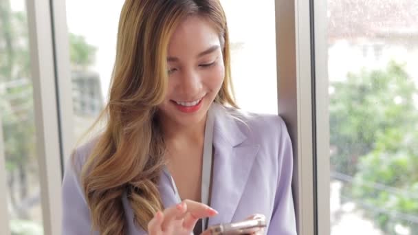 Όμορφη Ασιάτισσα επιχειρηματίας Κρατώντας ένα smartphone, κουβεντιάζοντας online με τους πελάτες. Στεκόταν στο παράθυρο του γραφείου. Έννοια των γυναικών επιχειρηματιών. - Πλάνα, βίντεο