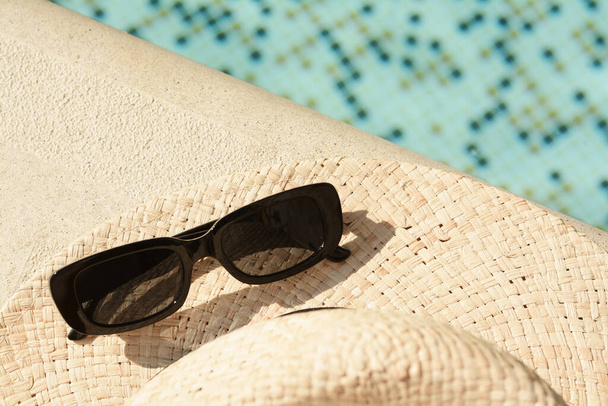 Stylish hat and sunglasses near outdoor swimming pool on sunny day. Space for text - Zdjęcie, obraz