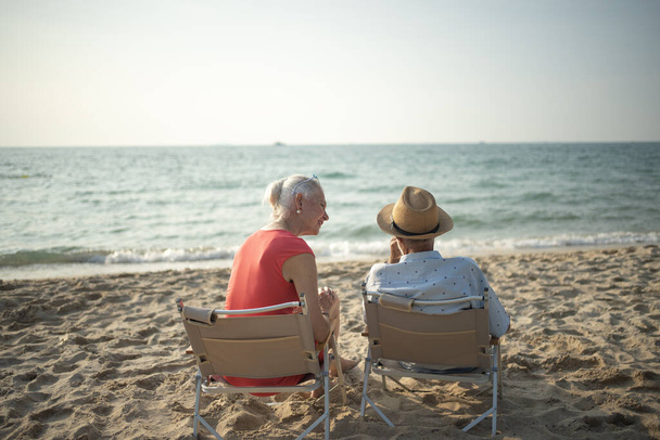Zwei ältere Männer und Frauen sitzen im Sessel am Strand und unterhalten sich, beobachten Sonne und Meer im Sommerurlaub und lächeln und genießen ihren Urlaub.. - Foto, Bild