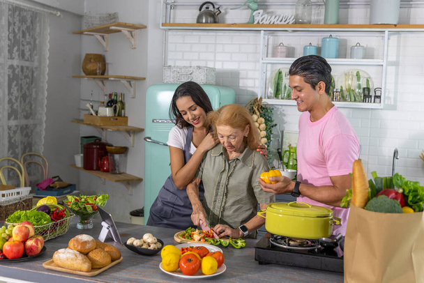 Jong stel werkt samen met grootmoeder om online kookcursus te volgen en thuis te koken. Gelukkig gezin kookt ontbijt met gezond eten samen - Foto, afbeelding