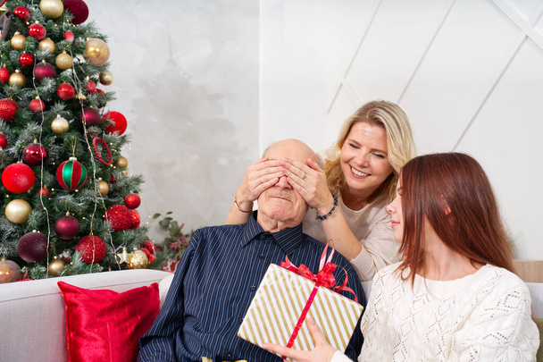 Az unokák ajándékot adnak nagyapának. Boldog nyugdíjas férfi ölelgeti az unokáját, miközben karácsonyi ajándékot kap otthon. Boldog család nagyapja aranyos izgatott unoka. - Fotó, kép