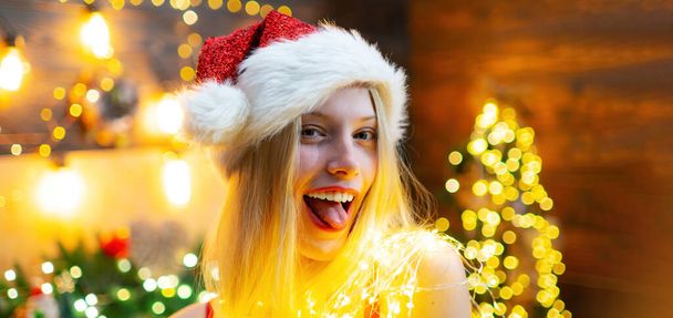Funny woman in Santa hat on christmas background. Close up portrait of sexy girl flirting, show tongue. Face of young smiling hipster woman on Christmas interior. Positive girl going crazy - Fotografie, Obrázek