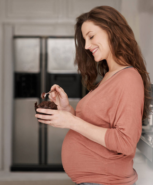 Poddaje się jej ciążowym zachciankom. młoda ciężarna kobieta jedząca babeczkę w kuchni - Zdjęcie, obraz