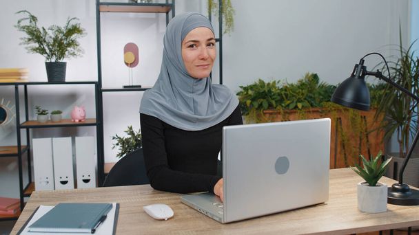 Молодая мусульманка-предпринимательница фрилансер в хиджабе открывает головной платок ноутбука начинает работать отправляет сообщения, делает онлайн покупки, смотрит фильмы. Арабская девушка использует ноутбук в современном рабочем пространстве домашнего офиса - Фото, изображение