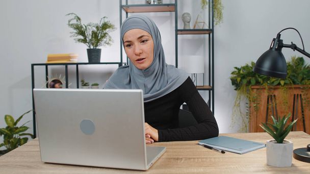 自宅のオフィスのデスクでラップトップコンピュータで働いているヒジャーブを身に着けているビジネス女性は、従業員のクライアント、上司とオンライン通信ビデオ通話について話します。ムスリム女子学生フリーランスカスタマーサポートサービス - 写真・画像