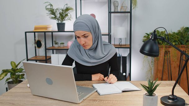 ラップトップコンピュータ上の自宅のオフィスでノートブックとヒジャーブの研究でイスラム教徒のビジネス女性は、ウェブカメラに話して会議ビデオ通話を行います。オンライン教師の電子リモートチャットで学ぶ。遠隔教育オンライン - 写真・画像