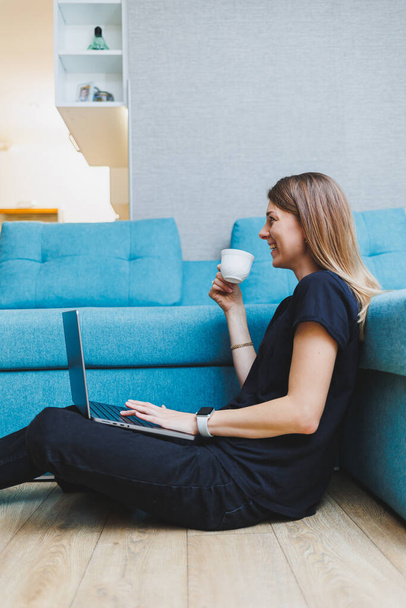 Mujer joven disfrutando del café de la mañana usando el ordenador portátil mientras está sentado en el sofá en casa, atractiva estudiante feliz escribiendo en el ordenador, divertirse en línea o charlar con amigos mientras se relaja en el sofá - Foto, Imagen