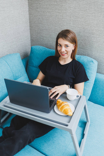 Приваблива молода жінка використовує ноутбук, сидячи на дивані вдома з кавою і круасанами. Робота в Інтернеті на відстані
. - Фото, зображення
