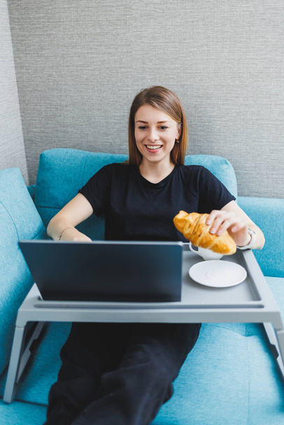 Ελκυστική νεαρή γυναίκα που χρησιμοποιεί φορητό υπολογιστή, ενώ κάθεται στον καναπέ στο σπίτι με καφέ και κρουασάν εργασίας. Εργασία σε απευθείας σύνδεση σε απόσταση. - Φωτογραφία, εικόνα