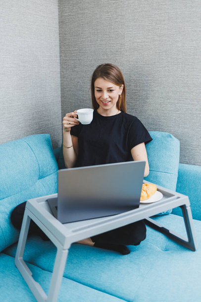 Młoda kobieta korzystająca z porannej kawy przy użyciu laptopa siedząc na kanapie w domu, atrakcyjna szczęśliwa studentka pisząca na komputerze, bawiąca się online lub rozmawiająca z przyjaciółmi relaksując się na kanapie - Zdjęcie, obraz
