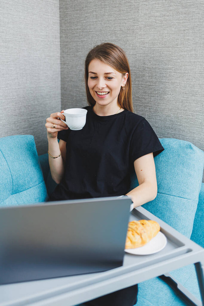 Junge Frau genießt Morgenkaffee mit Laptop, während sie zu Hause auf dem Sofa sitzt, attraktive, glückliche Studentin tippt am Computer, hat Spaß im Internet oder chattet mit Freunden, während sie es sich auf dem Sofa gemütlich macht - Foto, Bild