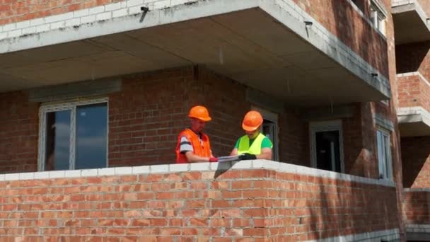Construction of a house. Engineers and architects review the construction plan. Two engineers wearing safety helmets are having a discussion on a construction site - Footage, Video