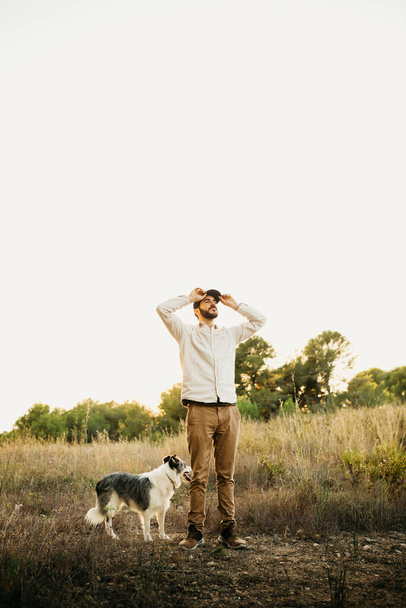 Portret van een jonge man met een baard met een zwarte pet, met zijn hond, op het platteland - Foto, afbeelding
