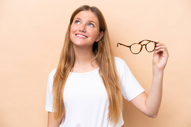 Young pretty blonde woman with glasses isolated on beige background looking up while smiling - 写真・画像
