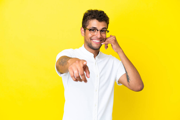 Young handsome Brazilian man isolated on yellow background making phone gesture and pointing front - Zdjęcie, obraz