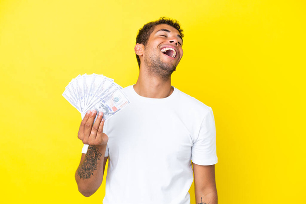 Mladý brazilský muž brát spoustu peněz izolovaných na žlutém pozadí se směje - Fotografie, Obrázek