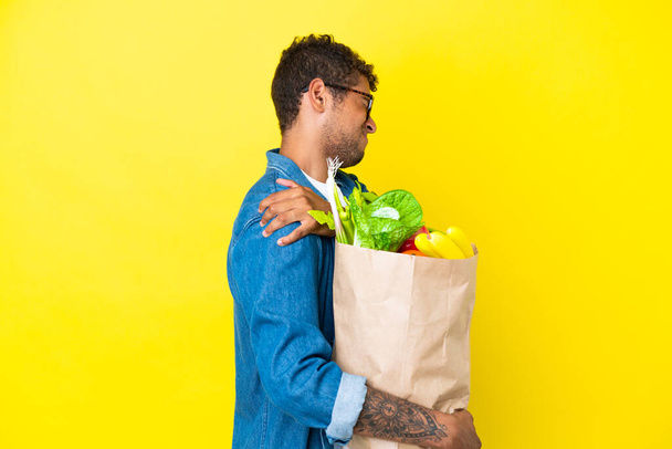 Young Brazilian man holding a grocery shopping bag isolated on yellow background suffering from pain in shoulder for having made an effort - Photo, image