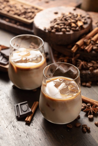 Ιρλανδική κρέμα και coffee cocktail σε ποτήρια με πάγο σε παλιό ξύλινο φόντο. Κόκκοι καφέ, κανέλα, γλυκάνισο, και τα κομμάτια της σοκολάτας είναι διάσπαρτα στο τραπέζι.  - Φωτογραφία, εικόνα